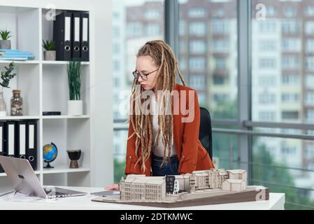 Experienced stylish female architect with dreadlocks working with cardboard model of residential area in architectural bureau Stock Photo