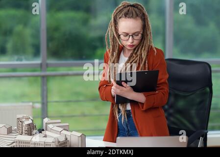 Adorable female engineer stands near constructed model and doing notes into the technical documents. Stock Photo