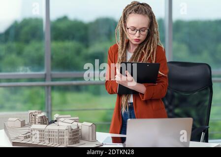 Adorable female engineer stands near constructed model and doing notes into the technical documents. Stock Photo