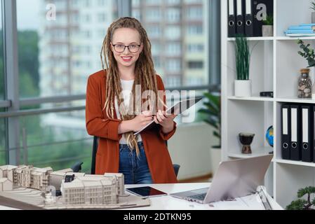 Adorable female engineer stands near constructed model and doing notes into the technical documents. Stock Photo