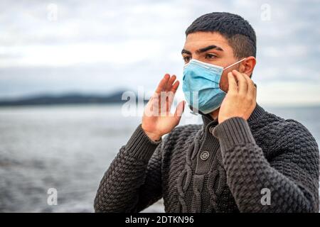 Handsome guy in dark wool sweater outdoor in sea resort has just worn face medical protection mask against Coronavirus pandemic virus - Young man livi Stock Photo