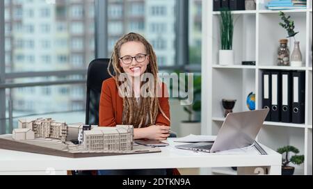 Stylish female architect in glasses with dreadlocks sits at her workplace with mock-up of future building. Stock Photo