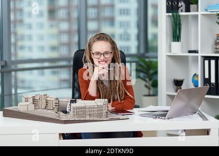 Stylish female architect in glasses with dreadlocks sits at her workplace with mock-up of future building. Stock Photo