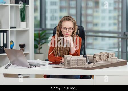 Stylish female architect in glasses with dreadlocks sits at her workplace with mock-up of future building. Stock Photo
