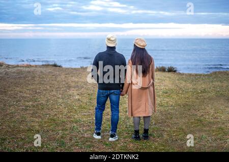 Romance scene of young heterosexual couple view from back holding hands watching the horizon on the sea - Two unrecognizable people wearing fall-winte Stock Photo