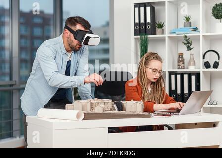Professional architect in augmented reality goggles working with mock up of building and woman coworker with laptop. Stock Photo
