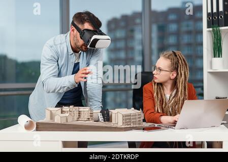 Professional architect in augmented reality goggles working with mock up of building and woman coworker with laptop. Stock Photo