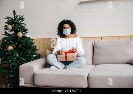 Millennial african american woman wearing a protective mask ah holdig box with red ribbon. Health protection during Coronavirus Christmas celebrating Stock Photo