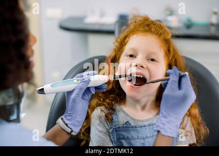 Close up view of little red haired girl patient having dental treatment at dentist's office. Young female african dentist making tooth restoration Stock Photo