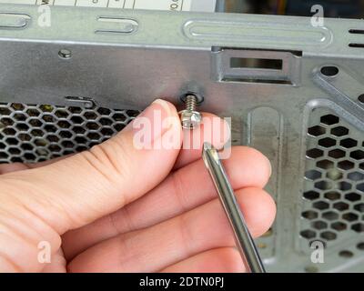 Woman fingers remove the screws holding the power supply unit in a tower case with a screwdriver. Personal computer PSU replasing, hardware equipment. Stock Photo