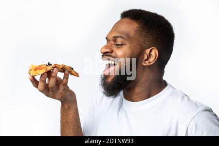 African Guy Eating Slice Of Pizza, White Background, Side-View Stock Photo