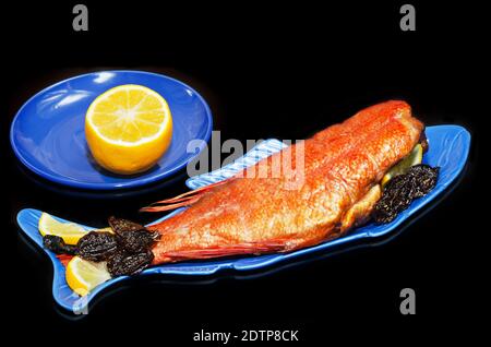 Hot smoked sea bass with lemon and dried spicy plums, close-up on a fish-shaped plate and black background. Seafood for proper nutrition Stock Photo