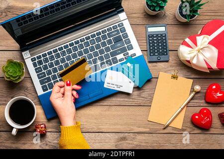 Girl holds debit card, chooses gifts, makes purchase, laptop, coffee cup, two hearts, bag on wooden table Top view Flat lay Holiday shopping list Stock Photo