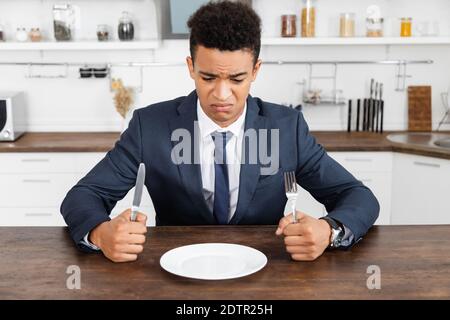 frustrated african american man holding cutlery and looking at empty plate Stock Photo