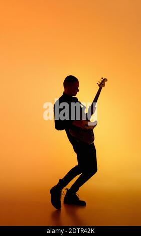 Party. Silhouette of young male guitarist isolated on orange gradient studio background in neon light. Beautiful shadow in action, performing. Concept of human emotions, expression, ad, music, art. Stock Photo