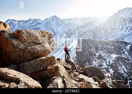 Man with backpack and red Santa Heat stands on the rock in the beautiful mountains with rising sun at background