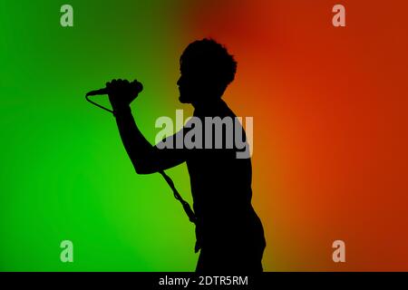 Voice. Silhouette of young male guitarist isolated on green-orange gradient studio background in neon. Beautiful shadow in action, performing. Concept of human emotions, expression, ad, music, art. Stock Photo