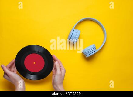 Women's hands hold vinyl record on yellow background with over-ear headphone. Pop culture, retro style, 80s. Minimalism. Top view. Flat lay Stock Photo