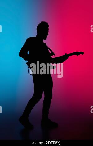 Party. Silhouette of young male guitarist isolated on blue-pink gradient studio background in neon. Beautiful shadow in action, performing. Concept of human emotions, expression, ad, music, art. Stock Photo