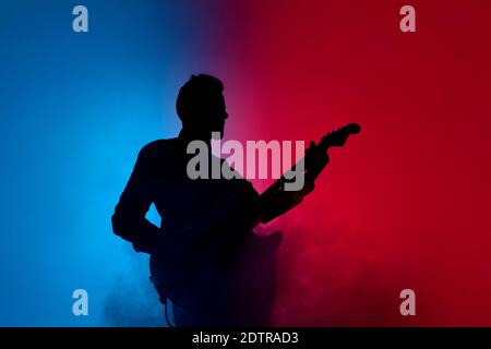 Smoke. Silhouette of young male guitarist isolated on blue-pink gradient studio background in neon. Beautiful shadow in action, performing. Concept of human emotions, expression, ad, music, art. Stock Photo