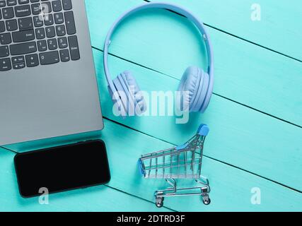 Online shopping. Buyer concept, consumer. Laptop, headphones, smartphone, toy shopping trolley on blue wooden background. Top view Stock Photo