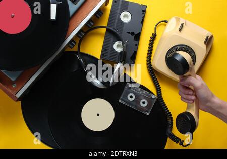 Retro attributes from the 80s on yellow background. Female hand holds the handset of vintage rotary phone against the background of vinyl player, vide Stock Photo