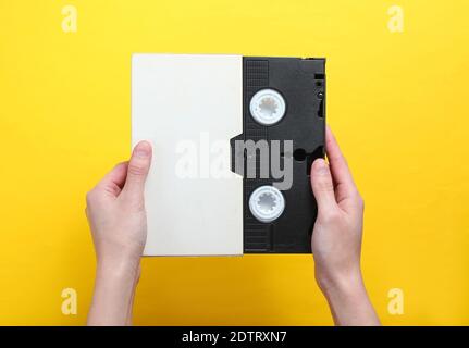 Woman hands holding videocassette in cover, videotape on a yellow background.Top view, minimalism. Stock Photo