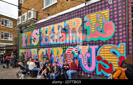 LONDON SPITALFIELDS BRICK LANE AREA LARGE WALL WITH ART AND PEOPLE EATING AND DRINKING Stock Photo