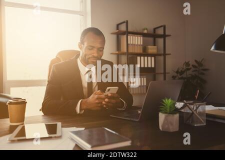 Portrait of smart afro american chief executive hr guy sit desk wait job applicant search social network news on smartphone wear suit tux tuxedo in Stock Photo