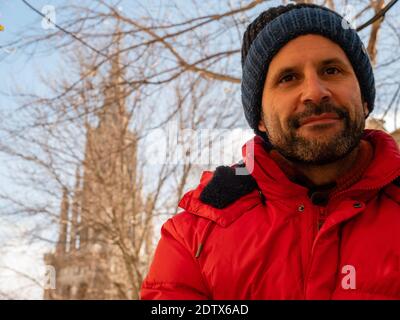 MAN IN RED JACKET AND BLUE HAT, SITTING WITH THE CATHEDRAL OF TOLEDO BEHIND HIM Stock Photo