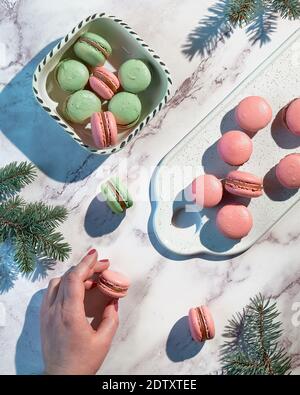 Pink and mint green macaroons on a tray and in ceramic bowl. Tasty creative dessert in green and red on marble table with female and winter fir twigs. Stock Photo