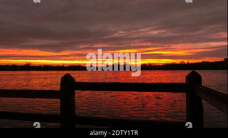 Amazing sunrise or sunset over a lake. Dramatic sky with clouds on the horizon with water and wooden fence on the foreground. Stock Photo