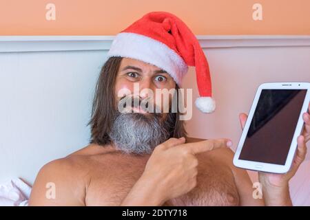 Close-up view of bearded with long hairs caucasian man in Santa Claus hat uses a tablet PC laying on the bed. He looks at the camera in surprise, pointing at something on the tablet. Holiday, festive and new year concept.  Stock Photo