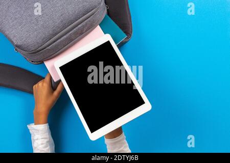 Kid hands taking digital tablet computer from gray backpack over light blue background. Top view Stock Photo