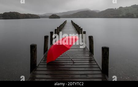 A rainy grey day on the shores of Coniston Water, English Lake District, UK. The red umbrella adds a splash of color to this almost monochrome photo. Stock Photo