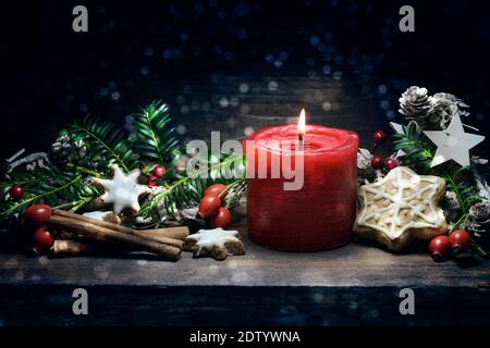 Red burning candle, gingerbread cookies, rose hips, branches and cones as Christmas or Advent decoration on a dark blue wooden background, copy space, Stock Photo