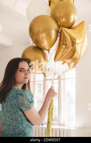 Young beautiful girl in blue dress with white polka dots celebrates her birthday and enjoys the golden balloons. Birthday alone at home during self-is Stock Photo