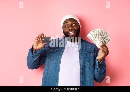 Dreamy african-american man showing credit card and dollars, thinking about shopping and smiling, standing over pink background Stock Photo