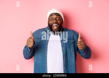 Impressed african-american hipster guy showing thumbs-up, approve and like offer, praising great choice, standing over pink background Stock Photo