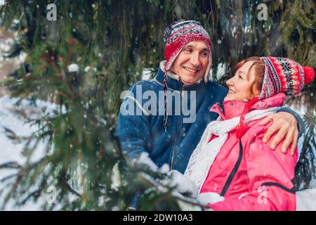 Senior couple hugging and laughing in winter forest. Happy man and woman walking outdoors under falling snow by fir tree Stock Photo
