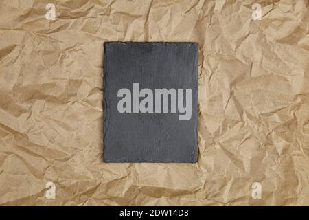 Slate board on crumpled craft paper background, top view. Black stone slate serving board, top view Stock Photo