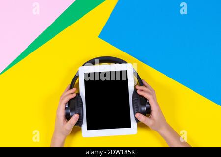Kid hands holding digital tablet with wireless headphones over blue pink and yellow background. Top view Stock Photo