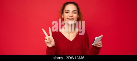 Photo of happy smiling woman with gathered curly hair wearing dark red sweater isolated over red background holding smartphone having fun and showing Stock Photo