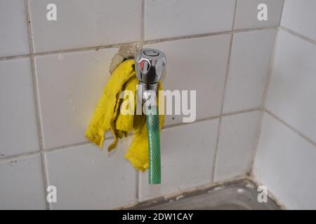 Metal faucet and piece of green hose on it. Yellow cleaning towel hanging on it with white faience background. Stock Photo