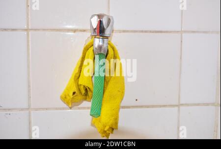 Metal faucet and piece of green hose on it. Yellow cleaning towel hanging on it with white faience background. Stock Photo