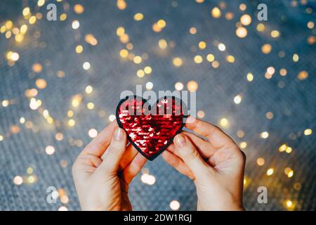 Red sequins heart in woman's hands on gray background with bokeh lights. Valentine's day and love concept. Stock Photo