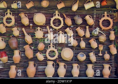 Rustic vintage clay jugs, pots and mugs hanging on wicker wall. Stock Photo