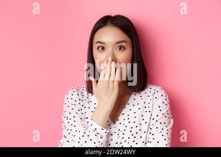 Image of shocked asian girl gossiping, gasping and cover mouth, stare at camera with complete disbelief, standing over pink background