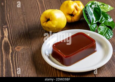 Homemade quince marmalade on the white plate, ripe yellow quince fruits with green leaves on the kitchen wooden table. Healthy traditional spanish des Stock Photo