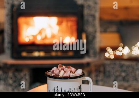 A mug of cocoa or hot chocolate with powdered marshmallows placed on a table by the fireplace Stock Photo